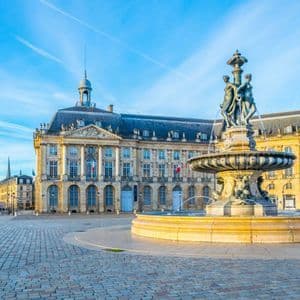 Une fontaine ornée de statues se dresse sur une vaste place pavée devant un grand bâtiment classique sous un ciel bleu.