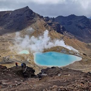 Un voyage de groupe WeRoad de randonneurs sur un sentier rocheux, surplombant deux lacs géothermiques bleus vifs d'où s'élève de la vapeur, dans une vallée volcanique.