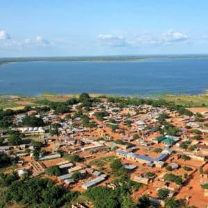 Luftaufnahme eines kleinen Dorfes mit unbefestigten Wegen und verstreuten Bäumen, gelegen am Ufer eines großen Gewässers unter blauem Himmel.