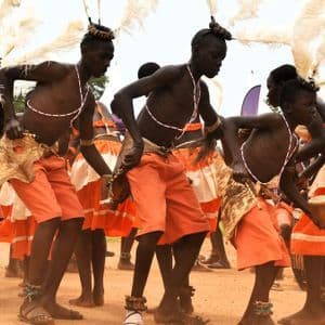Eine Gruppe von Menschen in orangefarbenen Trachten und weißen Federkopfschmuck führt einen kulturellen Tanz im Freien auf unbefestigtem Boden auf.