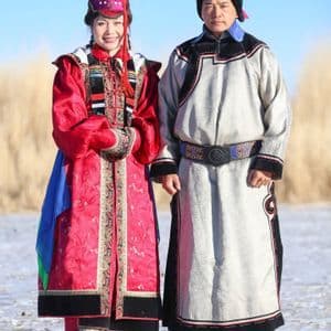 A man and a woman in colorful, traditional Mongolian attire stand side-by-side in a field under a clear blue sky.
