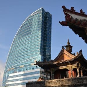 Un moderno grattacielo di vetro dal design curvo contrasta con il tetto decorato di un edificio tradizionale in primo piano, sotto un cielo azzurro.