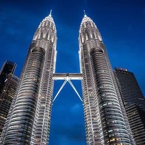 Zwei beleuchtete Zwillings-Wolkenkratzer mit einer verbindenden Skybridge, aus Froschperspektive vor einem dunkelblauen Nachthimmel.
