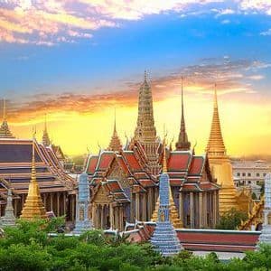 An ornate Thai temple complex with golden spires and tiled roofs against a colorful sunset sky.