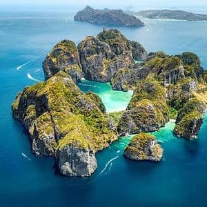 An aerial view of rocky, green-covered islands in a turquoise sea, with several small boats creating wakes in the water.