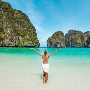 A woman seen from the back, wearing a white bikini and sarong, stands with arms outstretched in shallow turquoise water, facing large green cliffs across the bay.