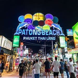 A large, colorful neon sign for a beach destination arching over a crowded street filled with people at night.