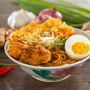 A bowl of curry noodle soup with chicken, tofu, and a hard-boiled egg, surrounded by fresh ingredients like chili peppers, ginger, and garlic.