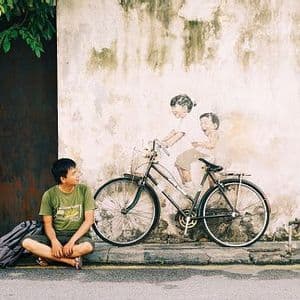 A young man sits on a sidewalk looking at a real bicycle parked in front of a mural of two children riding it.