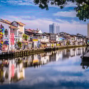 Colorful buildings, one with a large mural, line a calm river, their facades reflected in the water under a blue sky.