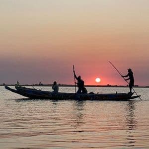 Siluetas de tres personas en un bote en el agua, con dos usando palos mientras el sol se pone en el horizonte.