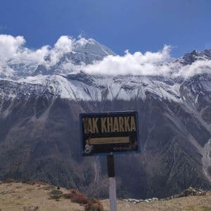 Un panneau "Yak Kharka" se dresse devant une vaste chaîne de montagnes enneigées sous un ciel bleu, avec un petit village dans la vallée en contrebas.