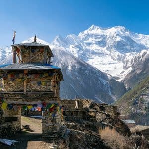 Un portail en pierre orné de drapeaux de prière colorés se dresse dans un village de montagne, avec d'imposants sommets enneigés en arrière-plan.