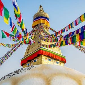 The golden spire of a Buddhist stupa, featuring painted eyes and adorned with colorful prayer flags against a clear blue sky.