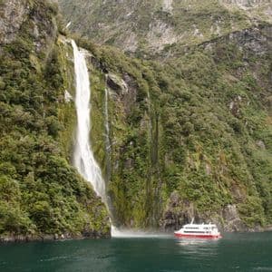 Ein rot-weißes Ausflugsboot fährt auf dem Wasser am Fuße eines hohen Wasserfalls an einer üppigen, felsigen Klippe.