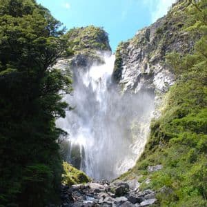 A powerful waterfall rushes down a rocky cliff, framed by lush green trees and vegetation in a steep gorge.