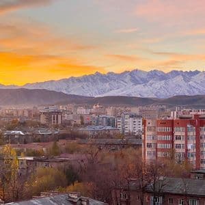 Un paesaggio urbano con numerosi edifici e alberi autunnali, sullo sfondo di una catena montuosa innevata sotto un tramonto dorato.