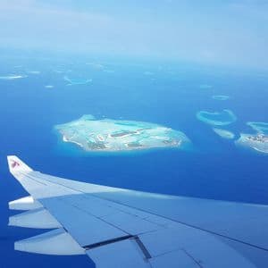 Une vue aérienne depuis la fenêtre d'un avion montrant l'aile au-dessus d'un océan d'un bleu profond parsemé de petites îles tropicales et d'atolls.