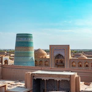 Uno scenario urbano con un grande e ornato minareto dalle piastrelle turchesi che si innalza sopra edifici e cupole color sabbia, sotto un cielo azzurro e limpido.