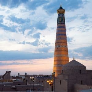 A tall, ornate minaret with blue and orange tiles rises above a cityscape of adobe buildings at sunset under a cloudy sky.