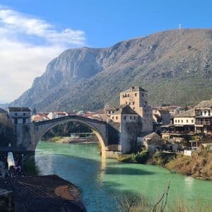 Eine Steinbogenbrücke überspannt einen türkisfarbenen Fluss, der durch eine Stadt mit Steingebäuden und Türmen fließt, mit einem großen Berg im Hintergrund.