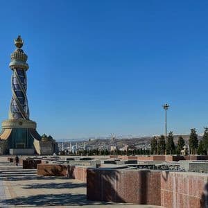 Un monumento ornato e a spirale con una cima dorata si erge in un'ampia piazza, con montagne innevate visibili in lontananza.