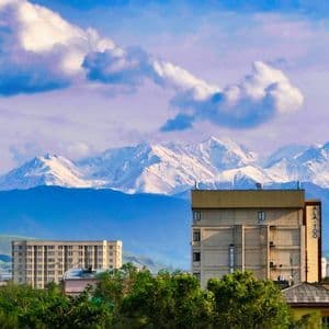 Uno skyline cittadino con edifici e alberi si trova di fronte a un'imponente catena montuosa innevata sotto un cielo azzurro nuvoloso.