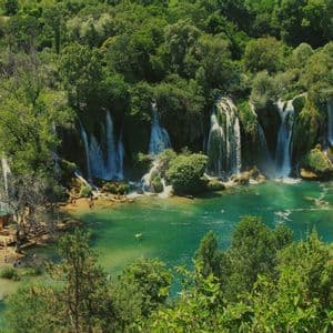 Breite Wasserfälle stürzen in einen türkisfarbenen Pool, in dem Menschen schwimmen, umgeben von dichtem grünem Wald.
