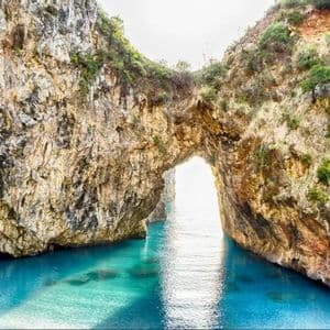 Un grande arco naturale di roccia si erge su acque limpide e turchesi, con il sole che splende luminoso attraverso l'apertura.