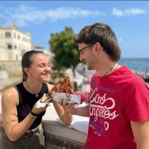 Un uomo con una camicia rossa offre una fetta di pizza a una donna sorridente su una terrazza all'aperto con il mare sullo sfondo.