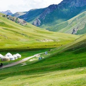 Un viaggio di gruppo WeRoad a cavallo su un sentiero sterrato accanto a un campo di yurte, immerso in una vasta e verde valle di montagna.