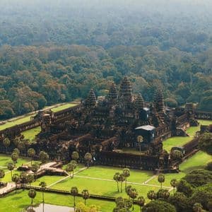 Una vista aerea di un grande e antico complesso di templi in pietra con torri, circondato da prati verdi, palme e una vasta giungla.