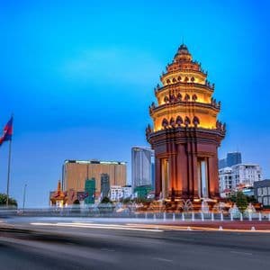 Ein reich verziertes, beleuchtetes Denkmal, umgeben von Springbrunnen in einer Stadt bei Dämmerung, mit Lichtspuren vom Verkehr im Vordergrund.
