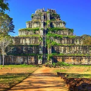 Un sentiero sterrato porta a un grande e antico tempio piramidale in pietra, ricoperto di vegetazione verde, sotto un cielo azzurro e limpido.