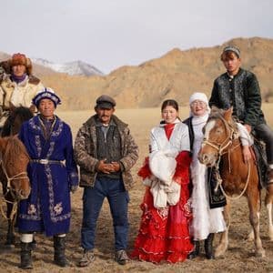 Un gruppo di persone in abiti tradizionali posa per una foto con cavalli e un'aquila reale in un vasto paesaggio collinare.