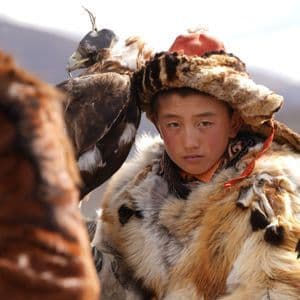 A young boy in traditional fur clothing and hat stands next to a hooded golden eagle, looking directly at the camera.