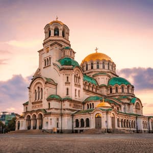 Una gran catedral blanca con cúpulas verdes y doradas se alza en una plaza adoquinada bajo un cielo de atardecer colorido.