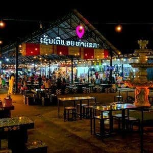 Un marché nocturne en plein air avec une fontaine décorative au premier plan, rempli de tables et d'étals sous des guirlandes de lumières chaudes.