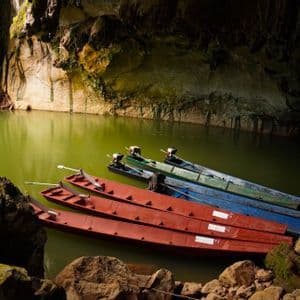 Plusieurs bateaux à longue queue rouges, bleus et verts sont amarrés sur une eau vert trouble, à l'intérieur d'une grotte rocheuse.