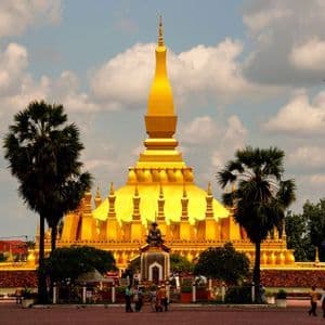 Un grand stupa doré avec une haute flèche centrale se dresse dans une cour, flanqué de palmiers sous un ciel partiellement nuageux.