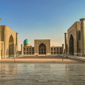 A large, empty plaza surrounded by ornate, tiled buildings with towering pillars and blue domes under a clear sky.