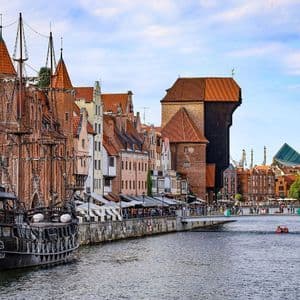 Un barco negro estilo pirata está atracado en un río frente a una ciudad con arquitectura histórica de ladrillo y una grúa portuaria medieval.
