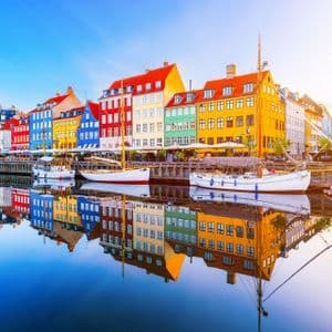 Una fila di edifici colorati e diverse barche si riflettono nelle acque calme di un porto sotto un cielo azzurro e limpido.