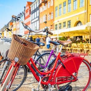 Una bicicletta rossa con un cestino di vimini è parcheggiata su una strada di ciottoli davanti a edifici colorati e un caffè all'aperto.