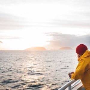 Una persona con un impermeabile giallo e un berretto rosso si appoggia a una ringhiera, guardando il mare e le isole lontane al tramonto.