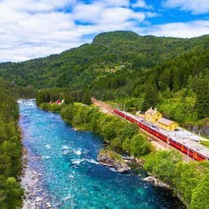 Una veduta aerea di un treno rosso in stazione, costeggiando un fiume turchese che attraversa una valle montana lussureggiante e boscosa.