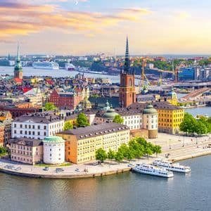 Veduta aerea di una storica città europea sull'acqua, con edifici colorati, una guglia di chiesa e barche ormeggiate lungo la riva al tramonto.