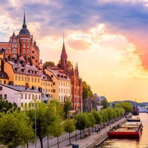 Edifici storici colorati con guglie e tetti scuri fiancheggiano una passeggiata lungofiume accanto a un ampio fiume al tramonto.