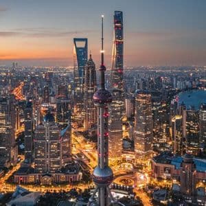 Lo skyline illuminato di una città moderna con numerosi grattacieli e un'imponente torre, vista dall'alto al crepuscolo.