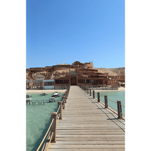 Un lungo molo di legno si estende su acque turchesi e limpide verso un grande resort balneare in legno costruito su un versante sabbioso sotto un cielo azzurro e sereno.